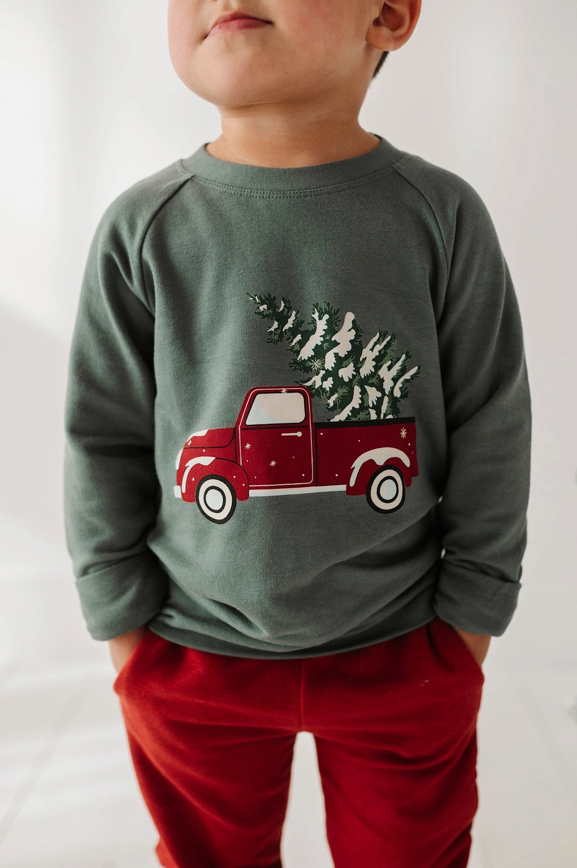 Child wearing a green sweatshirt with a red truck and Christmas tree design on a white background
