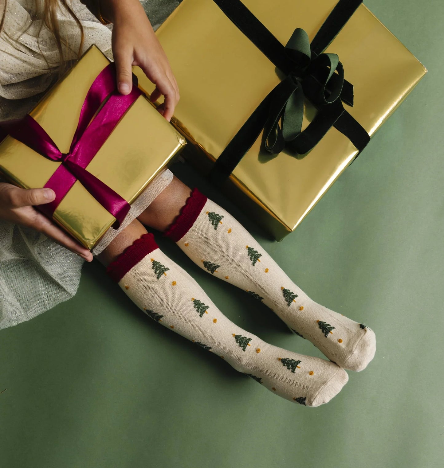 Person wearing knee-high socks with Christmas tree pattern, holding gold gift box on green background