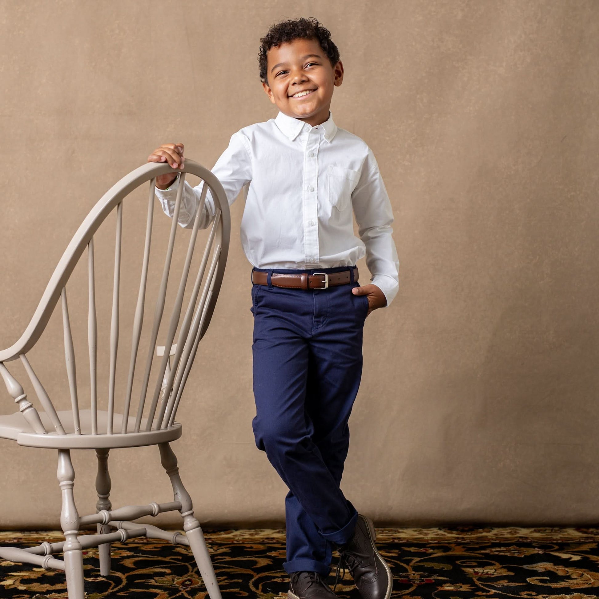 Young boy in a white shirt and blue pants standing next to a chair against a beige wall.
