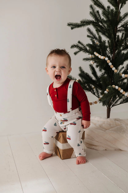 Child in a red shirt and patterned overalls standing in front of a Christmas tree.