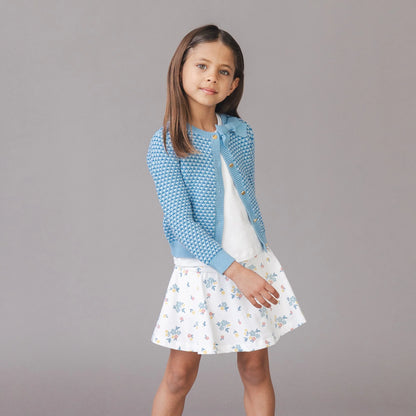 Young girl wearing a blue textured cardigan over a white dress with floral pattern on a gray background