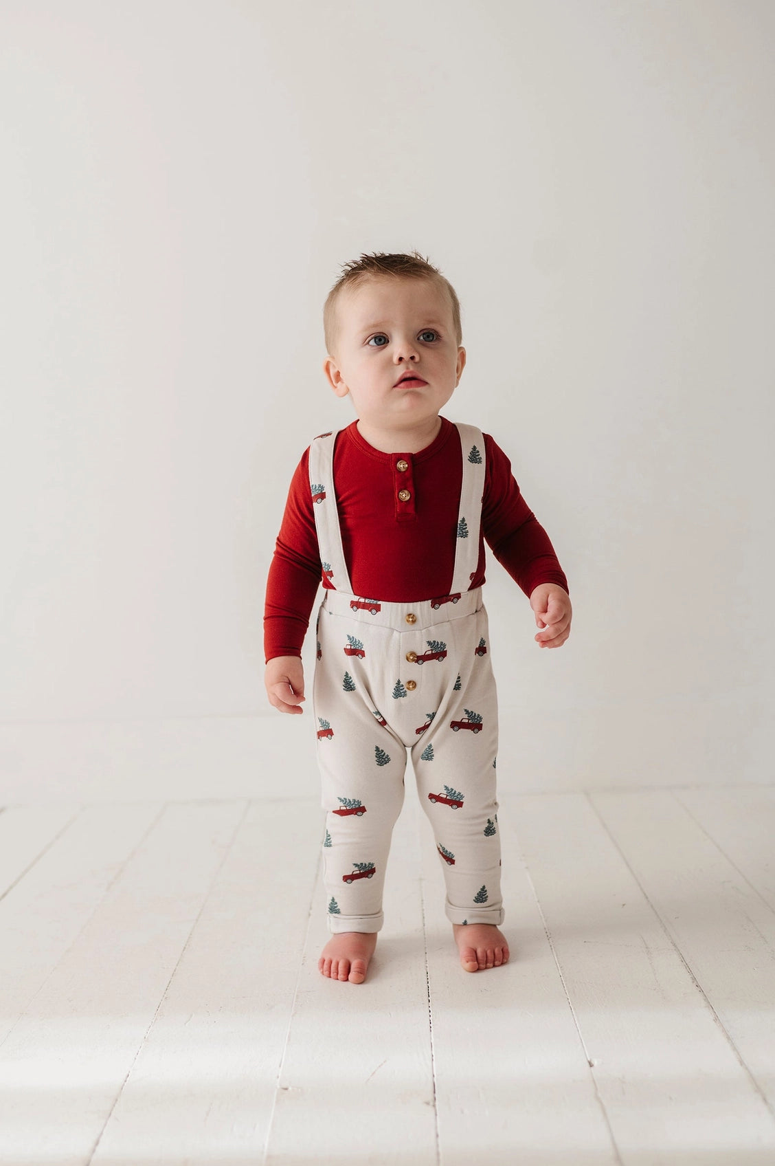 Baby wearing a red shirt and white overalls with vehicle pattern on a light background