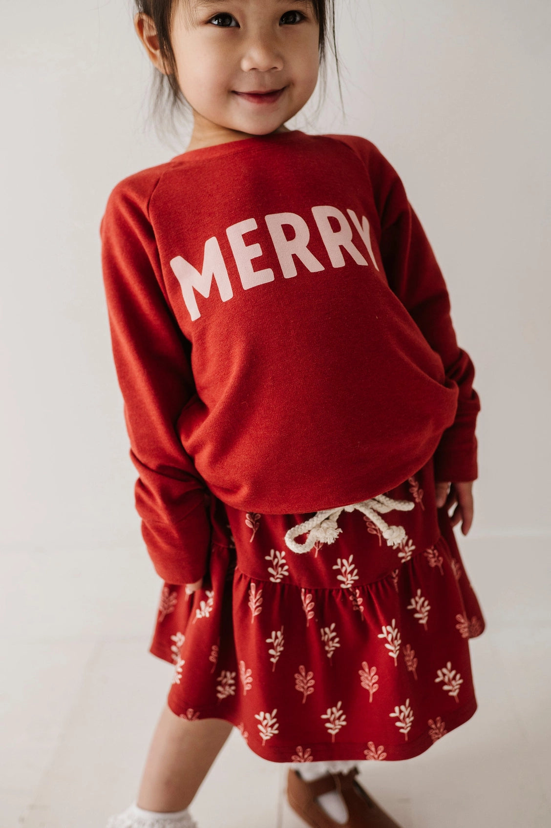 Child wearing a red sweater with 'MERRY' and a matching skirt with white patterns on a plain background