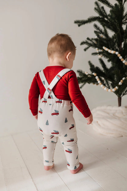 Child wearing a red shirt and patterned overalls standing in front of a Christmas tree.