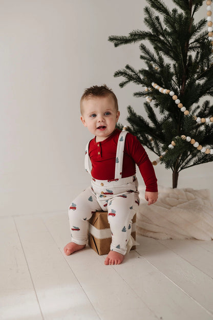 Baby in a red shirt and patterned pants sitting on a box in front of a Christmas tree.
